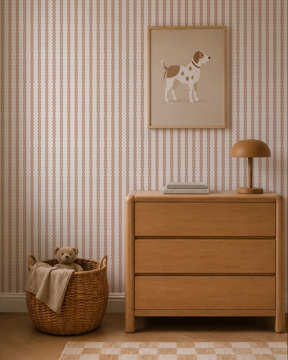 A cozy nursery with a wooden dresser, brown lamp, dog illustration, and a woven basket with a teddy bear and beige blanket, featuring Little Abacus Stripe Wallpaper in Rose.