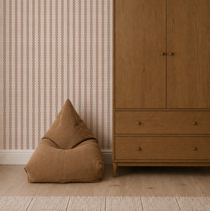 A brown bean bag sits next to a wooden wardrobe on a light wood floor, with Little Abacus Stripe Wallpaper in Rose decorating the background and a beige rug in the foreground.