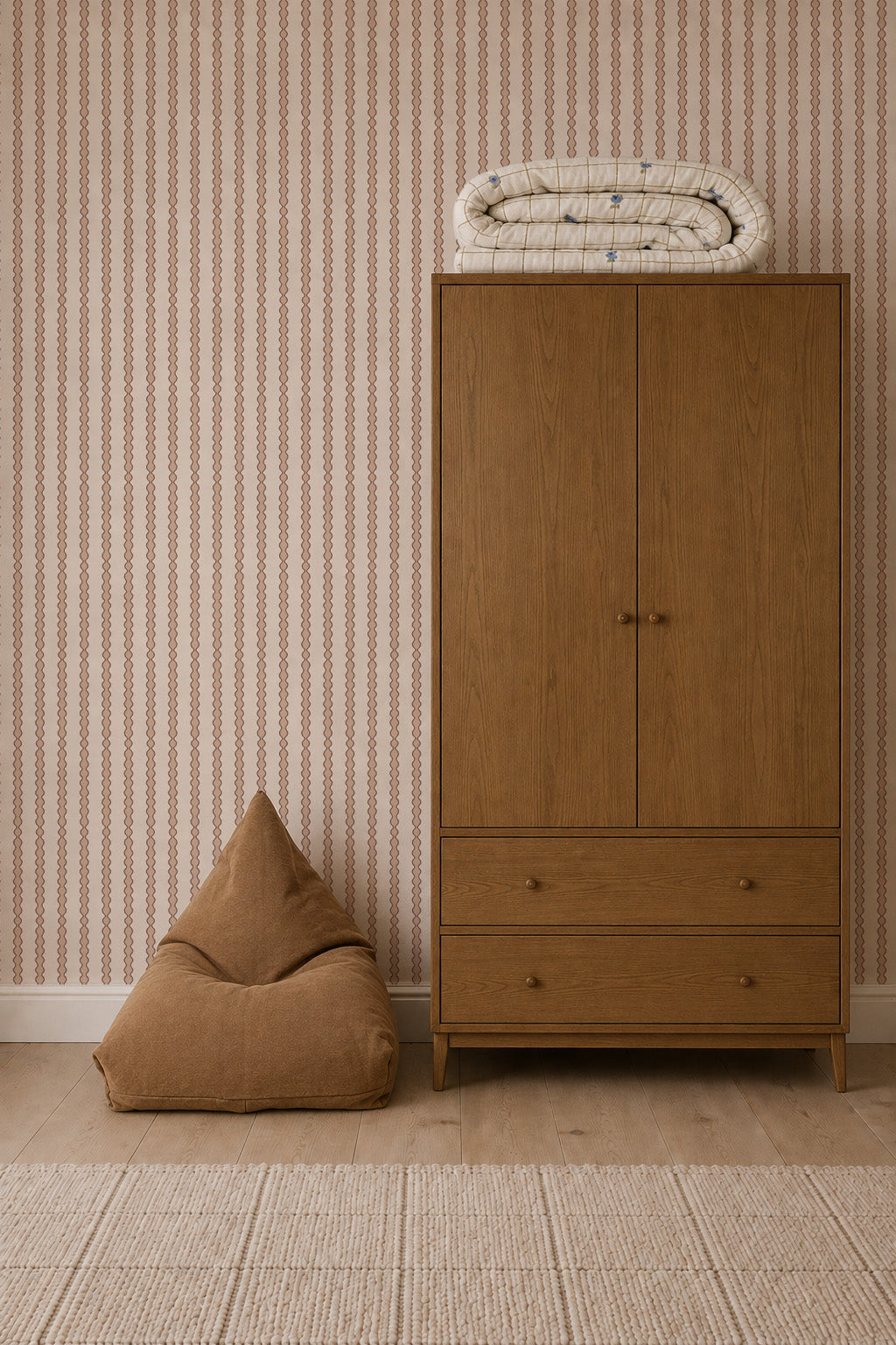 A wooden wardrobe stands beside a brown bean bag chair, with folded bedding on top. The room features Little Abacus Stripe Wallpaper in Rose, an elegant wall pattern, along with a woven rug and light wood flooring.