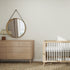 A minimalist nursery pairs a light wood crib, matching dresser, round mirror, lamp, and textured rug with Little Abacus Stripe Off White and Taupe Wallpaper Sample for a calm, neutral look. Ideal for wallpaper inspiration from online imagery.
