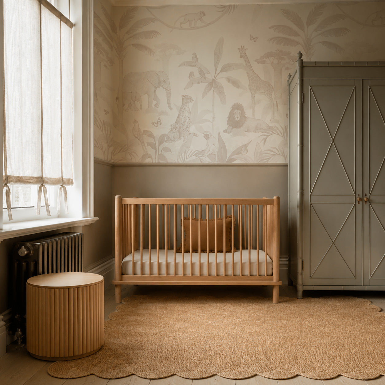 Neutral nursery with a wooden crib, round side table, scalloped rug, and a gray wardrobe. The wall features the African Safari Mural, Neutral—a softly toned wallpaper mural—while sunlight filters in through shaded windows.