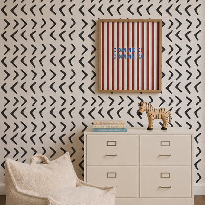 A beige dresser with books and a toy zebra on top stands against a wall adorned with Andy Arrow Wallpaper in Charcoal, which features striking black and white chevron patterns. Above the dresser hangs a red-and-white striped art piece with blue text. A cream-colored plush chair is in the foreground.