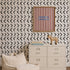 A beige dresser with books and a toy zebra on top stands against a wall adorned with Andy Arrow Wallpaper in Charcoal, which features striking black and white chevron patterns. Above the dresser hangs a red-and-white striped art piece with blue text. A cream-colored plush chair is in the foreground.