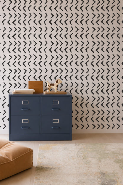 A blue four-drawer cabinet sits against a wall adorned with Andy Arrow Wallpaper in Charcoal, featuring a bold black and white chevron pattern. Decorative items and books are arranged on top of the cabinet, while a tan cushion and a partial rug rest on the light wood floor.