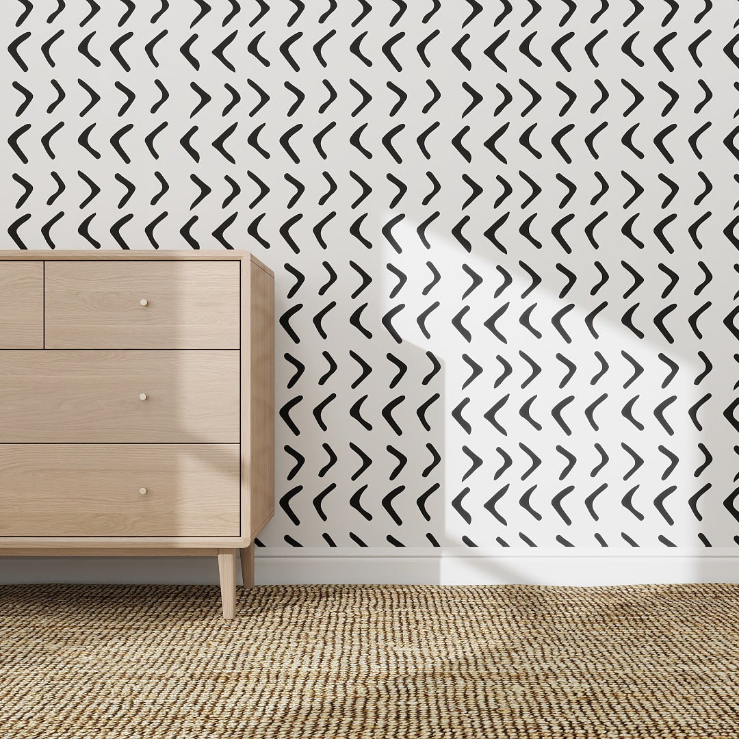 A light wood dresser sits on a woven rug in front of a white wall covered with Andy Arrow Wallpaper, Charcoal. Sunlight casts shadows over the bold black and white dash pattern and across the floor.