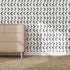 A light wood dresser sits on a woven rug in front of a white wall covered with Andy Arrow Wallpaper, Charcoal. Sunlight casts shadows over the bold black and white dash pattern and across the floor.