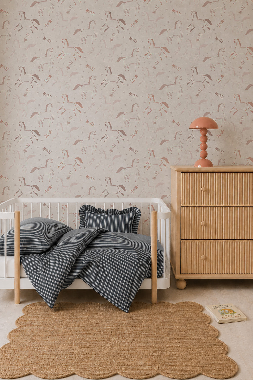A cozy nursery features a white crib with navy striped bedding, a scalloped jute rug, a rattan dresser with a coral lamp, and whimsical Astrid Unicorn Wallpaper adorned with abstract animal and nature motifs. A book lies on the floor nearby.