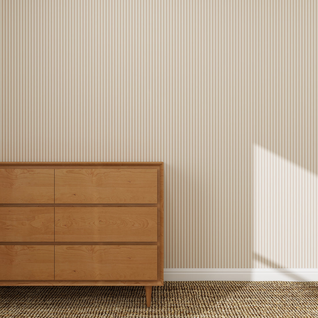 A wooden dresser with three drawers sits on a textured woven rug against a wall featuring Pico Stripe Wallpaper Rose | Sample, highlighting its beige and white stripes. Sunlight casts a shadow on the lower right side of the wall.