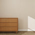 A wooden dresser with three drawers sits on a textured woven rug against a wall featuring Pico Stripe Wallpaper Rose | Sample, highlighting its beige and white stripes. Sunlight casts a shadow on the lower right side of the wall.