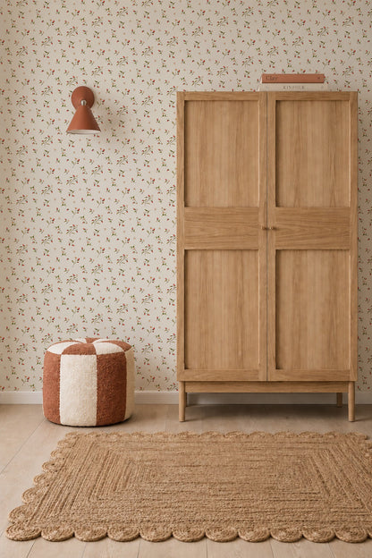 A cozy room featuring a light wood wardrobe with a book on top, a two-toned round pouf, textured natural fiber rug, wall sconce, and Berry and Bramble Wallpaper in soft botanical tones.