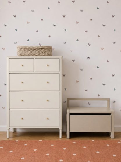 A cream-colored dresser and a small bench sit on a wooden floor. The wall behind is adorned with Bibi Butterflies Wallpaper, featuring a charming hand-drawn butterfly repeat pattern, while an orange rug with white dots is partially visible in the foreground.