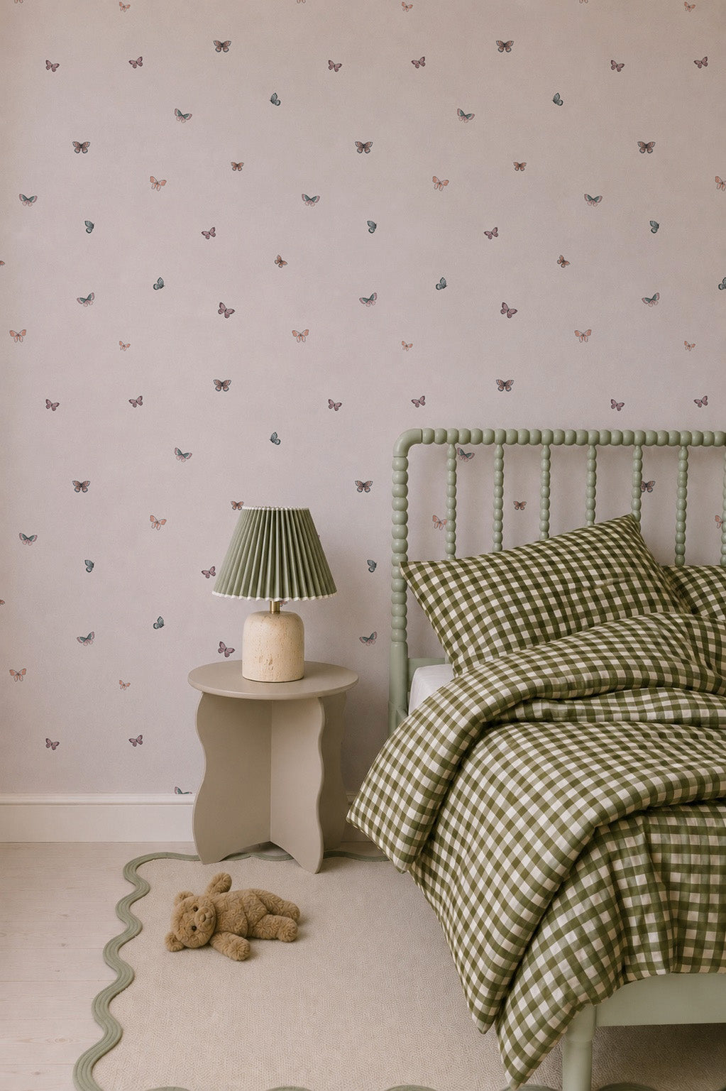 A cozy bedroom featuring the Bibi Butterflies Wallpaper with its delicate hand-drawn butterfly repeat pattern, a green checkered bed with matching pillows, a teddy bear on the floor, and a small table topped with a lamp.
