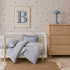 A cozy nursery features a white crib, blue-striped bedding, a wooden dresser, a blue lamp, books scattered on the floor, and charming Bibi Butterflies Wallpaper in a hand-drawn repeat pattern. A framed letter B hangs above the crib.