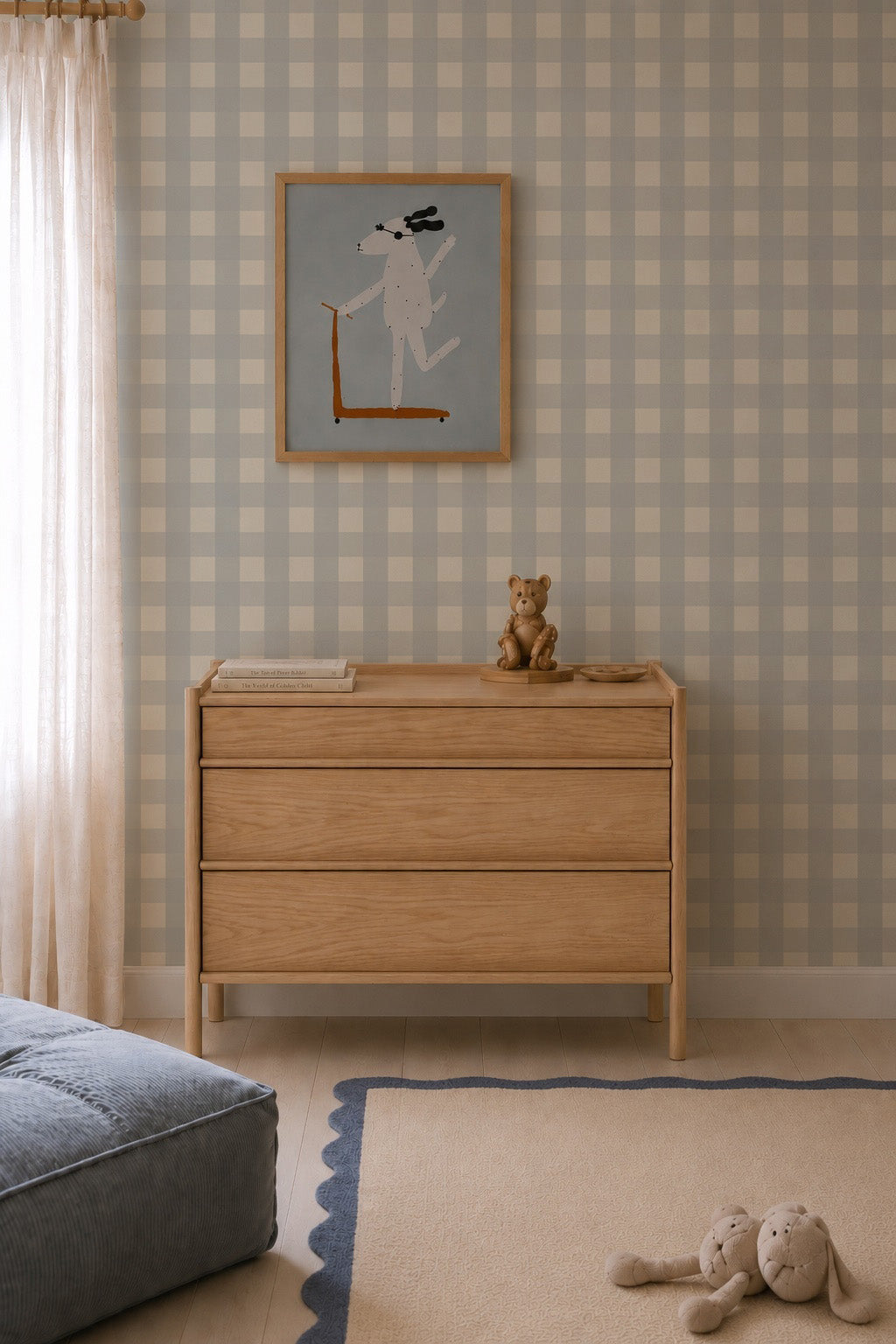 A wooden three-drawer dresser against Large Check Wallpaper, Blue, topped with a small teddy bear and books, with a framed dog illustration hanging above.