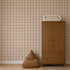 A wooden wardrobe stands against Large Check Wallpaper, Pink, with a folded patterned blanket on top and a brown pyramid-shaped beanbag on the floor.