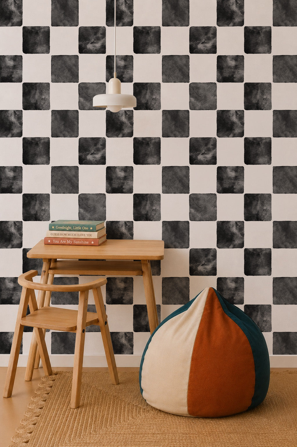 A small wooden desk and chair sit on a woven rug in front of Billys Checkered Wallpaper, Black. Books rest on the desk, a pendant lamp hangs above, and a round color-block beanbag completes this cozy, playful space.