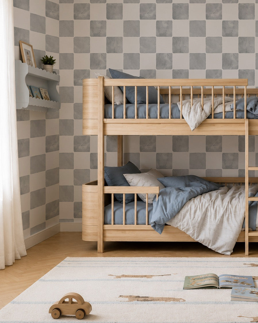 Children’s bedroom with wooden bunk beds, blue and white bedding, Billys Checkered Wallpaper in Blue, a shelf with toys and books, a striped rug, a wooden toy car on the floor, and an open book lying on the rug.