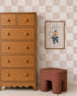 A wooden dresser with round knobs stands beside a small brown stool. Above it, a framed mouse illustration hangs on a wall decorated with Billys Checkered Wallpaper, Pink, adding charming wall decor.