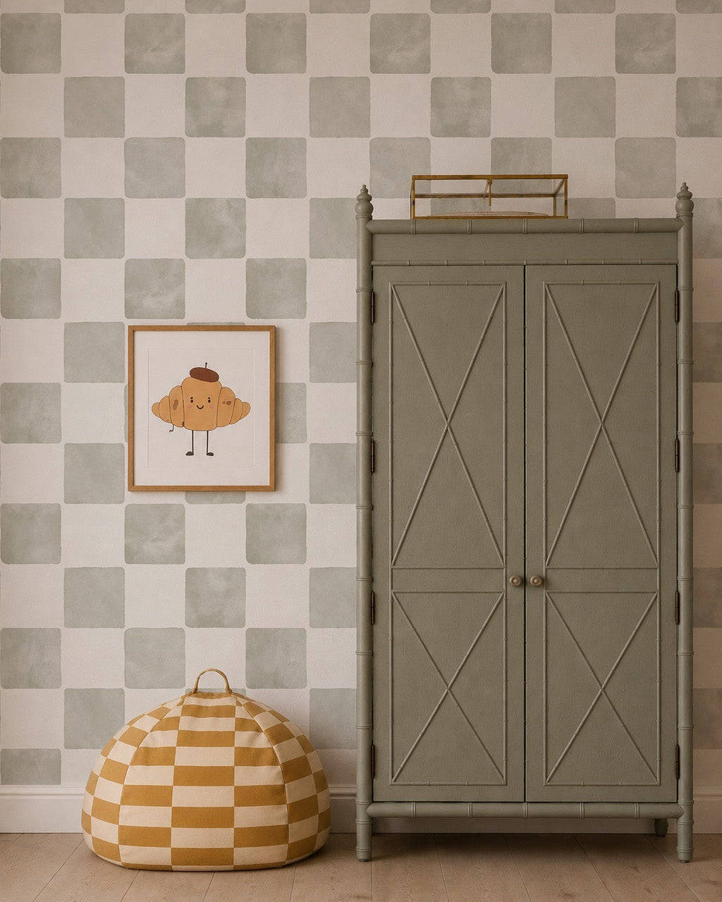 A gingham-patterned ottoman sits beside a tall olive-green armoire. Above, a framed cartoon croissant print hangs on a wall decorated with Billys Checkered Wallpaper in sage.
