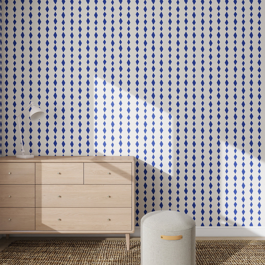 A minimalist room with a light wood dresser, white lamp, and round ottoman. Sunlight casts diagonal shadows on the wall adorned with Jolly Jester Wallpaper Repeat Pattern | Royal Blue. A woven rug covers the floor.