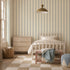 A cozy bedroom with a light wood bed, beige checkered rug, wooden dresser, and wicker laundry basket. The walls feature Scallop Stripe Wallpaper in Blue, and a blanket is draped over a bench at the foot of the bed.