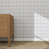 A wooden dresser stands on a woven carpet by a light gray wall decorated with the Checkered Wallpaper Repeat Pattern Blue | Sample, as sunlight casts a triangular shadow, highlighting the wallpapers quality.