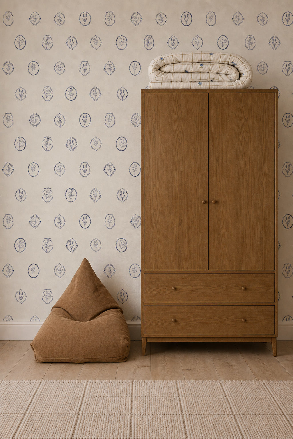 A brown bean bag sits beside a wooden wardrobe with two drawers, topped with folded blankets. The wall features Botanic Emblem Wallpaper, and a beige rug lies on the wooden floor.