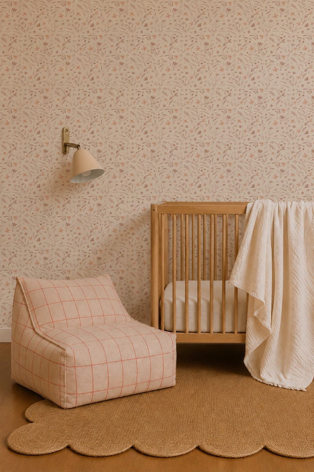 A cozy nursery with a wooden crib, a checkered chair, and a scalloped jute rug on wood floors. The wall features Butterfly Meadowland Wallpaper in Dune and a small wall lamp, adding gentle nature-inspired decor.
