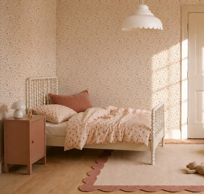 Cozy children’s bedroom with a white spindle bed, peach and cream bedding, matching pillow, pink nightstand with lamp, and a beige rug with scalloped pink border. The room features Butterfly Meadowland Wallpaper in Dune.
