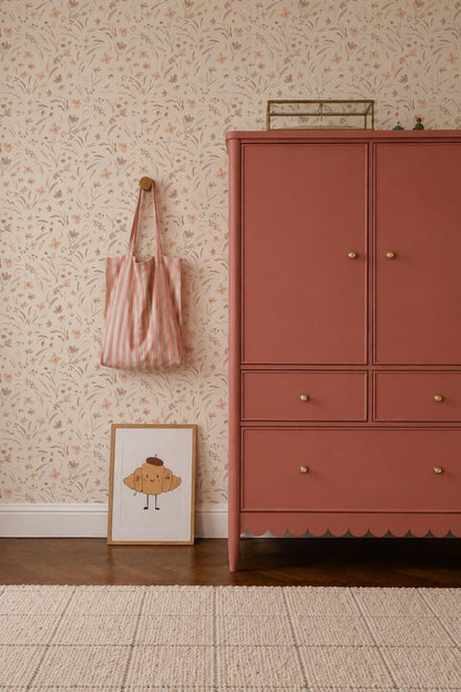 A dresser stands in a room with Butterfly Meadowland Wallpaper, Dune. A pink striped tote hangs on a wood peg, and a framed muffin illustration rests on the wooden floor beside a beige rug.