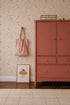 A dresser stands in a room with Butterfly Meadowland Wallpaper, Dune. A pink striped tote hangs on a wood peg, and a framed muffin illustration rests on the wooden floor beside a beige rug.