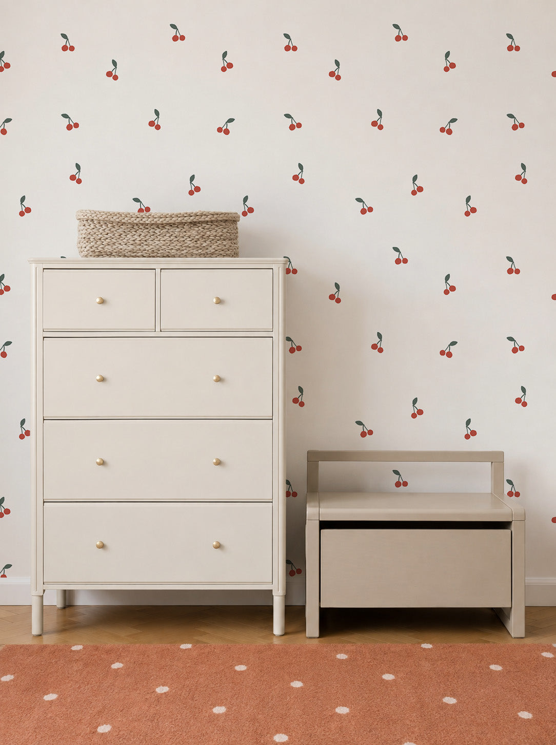 A light-colored dresser and bench sit against a wall adorned with Cherry Sprinkle Wallpaper, adding a playful touch to the space. A woven basket rests atop the dresser, while a rust-colored rug with white dots completes the cozy look.