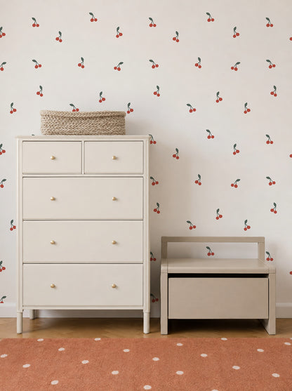 A light-colored dresser and bench sit against a wall adorned with Cherry Sprinkle Wallpaper, adding a playful touch to the space. A woven basket rests atop the dresser, while a rust-colored rug with white dots completes the cozy look.