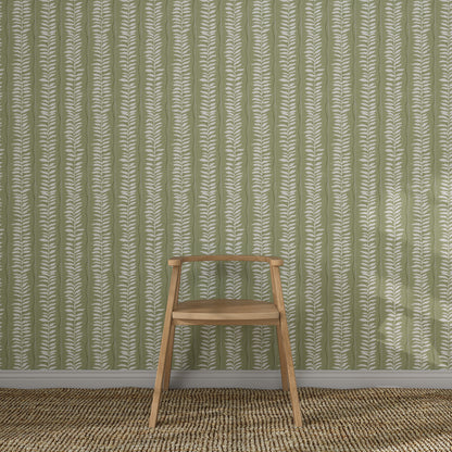 A wooden chair with a simple design sits on a woven rug in front of Meadow Leaf Stripe Wallpaper, Sage, its white botanical stripes adding charm to the cozy space.
