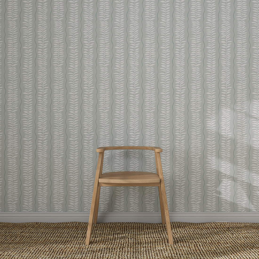 A simple wooden chair sits on a woven rug in front of Meadow Leaf Stripe Wallpaper, Sea Foam, featuring hand-painted vertical leaf designs in muted tones.