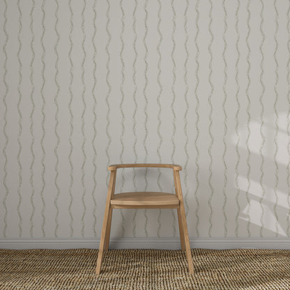 A light wooden chair with a curved backrest sits on a woven rug before a white wall featuring Ribbon Stripe Wallpaper in Sage. Sunlight casts partial shadows, creating an elegant children&