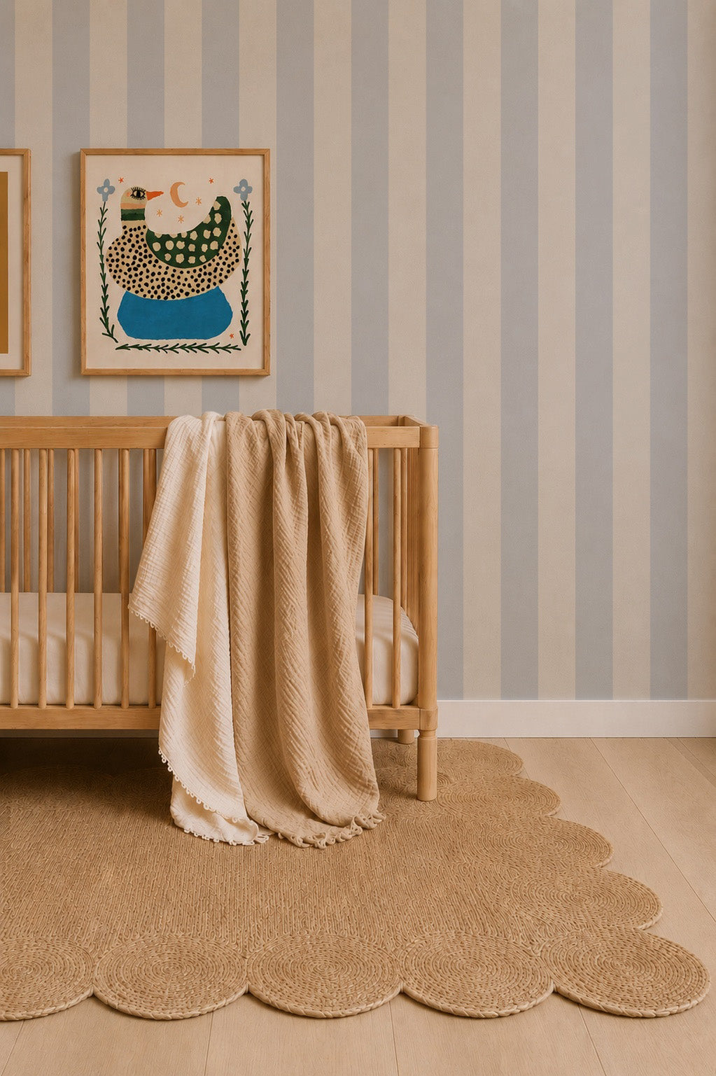 A minimalist nursery features a wooden crib, beige blanket, scalloped round rug, and Circus Stripe Wallpaper Sample in Sky. The soft hue pairs perfectly with two abstract animal prints above the crib.