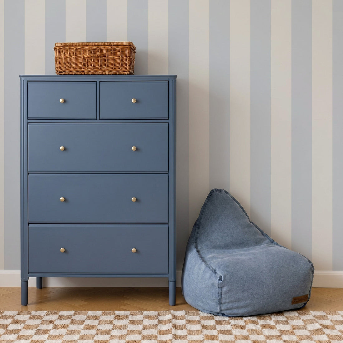 A blue dresser with four drawers stands by a blue bean bag chair. On top is a wicker basket. The wall features Circus Stripe Wallpaper Sample, Sky. A checkered beige and white rug covers the floor.