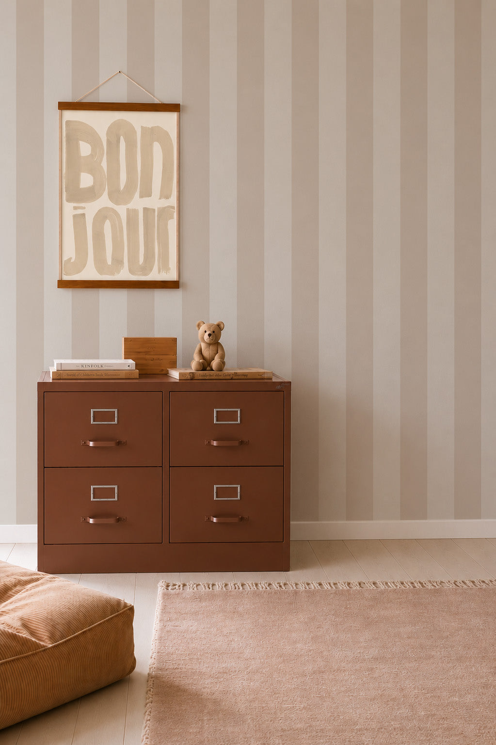A striped beige wall adorned with Circus Stripe Wallpaper, Soft Beige, sets a charming tone, while a BONJOUR sign hangs above a brown filing cabinet. A small teddy bear and books rest on the cabinet—perfect for little ones. A beige rug and pouf complete this cozy corner, ideal for nursery wallpaper inspiration.