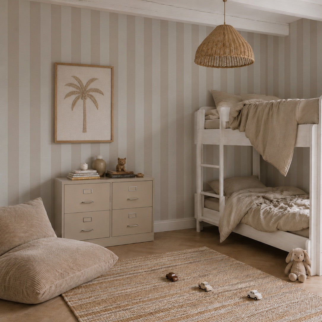 A cozy kids room features Circus Stripe Wallpaper, Soft Beige on the walls, complemented by white bunk beds, a wicker pendant lamp, a plush bean bag chair, a woven rug, a cabinet adorned with decorative items, and framed palm tree artwork. The neutral and minimalist decor showcases a perfect blend of style and playful charm thanks to the Circus Stripe Wallpaper.