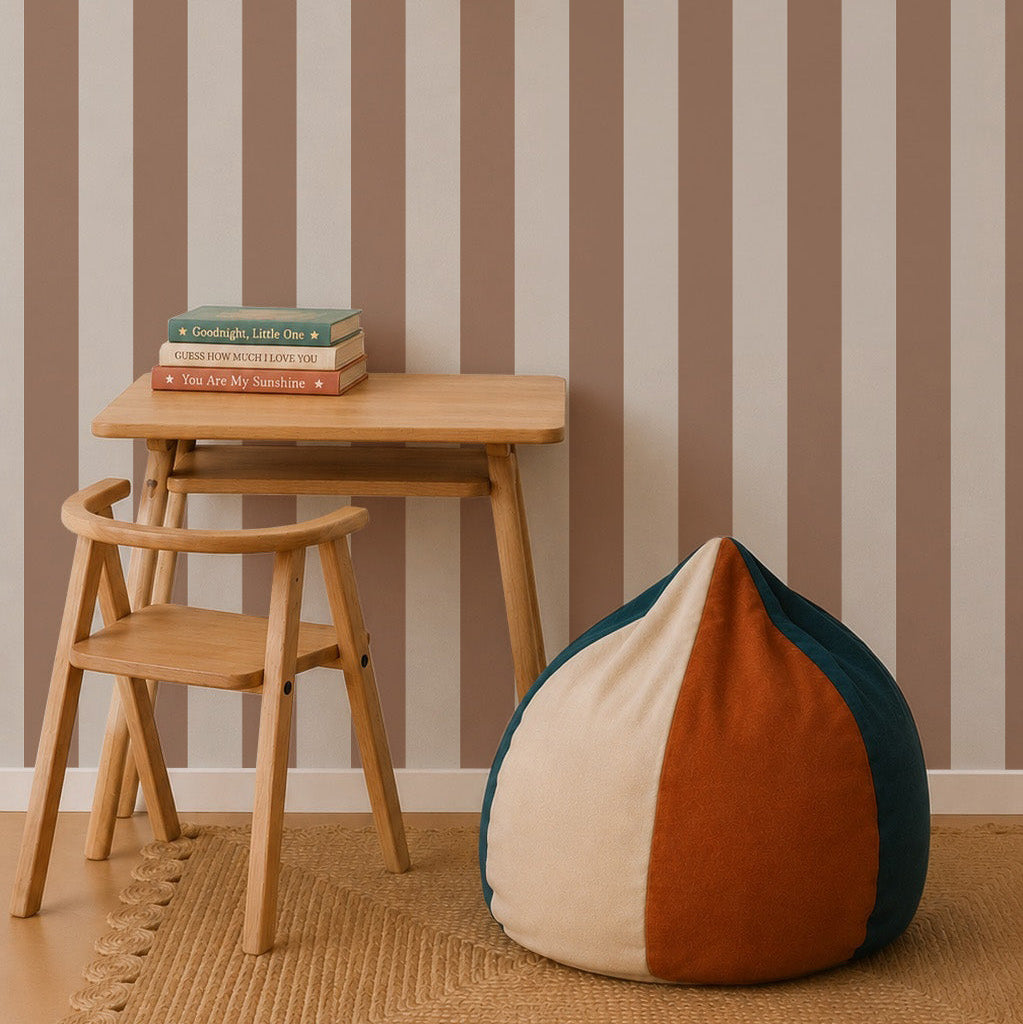 A small wooden desk and chair sit on a woven rug next to a color-blocked bean bag. Three books are stacked under a white pendant light, with vertical Circus Stripe Wallpaper, Latte in beige and brown as the background.