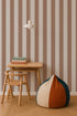 A small wooden desk and chair sit on a woven rug next to a color-blocked bean bag. Three books are stacked under a white pendant light, with vertical Circus Stripe Wallpaper, Latte in beige and brown as the background.