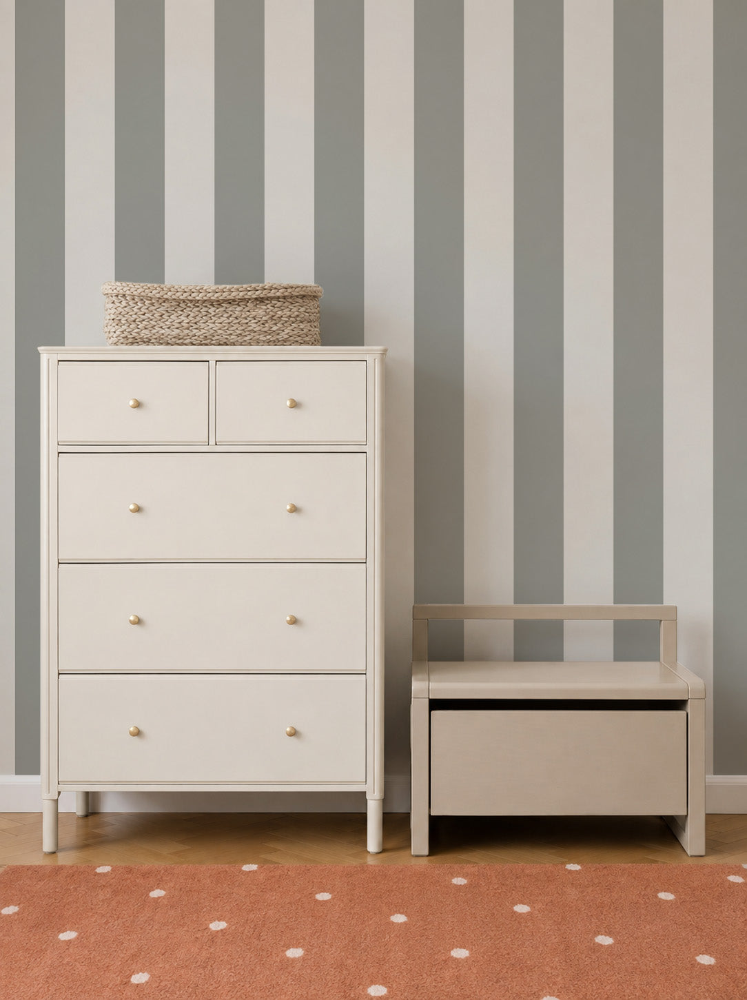 A cream-colored dresser and a small bench sit against a wall adorned with Circus Stripe Wallpaper in Matcha, featuring vertical gray and white stripes, on a floor with an orange polka-dot rug. A woven basket rests on top of the dresser.