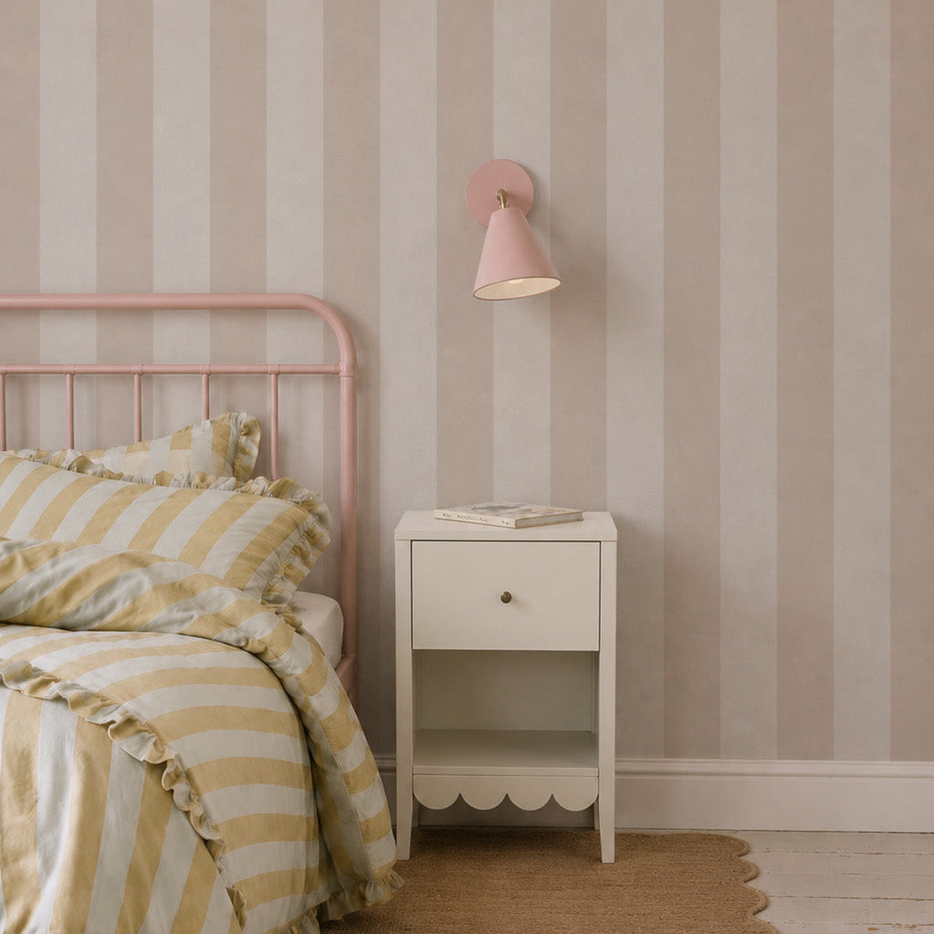 A bedroom featuring a pink metal bed, yellow and white striped bedding, a white nightstand with a book, and a pink wall lamp is brought to life by the playful charm of the Circus Stripe Wallpaper in Soft Beige.