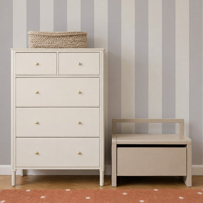 A cream-colored dresser and matching bench sit against a wall adorned with Circus Stripe Wallpaper, Stone, featuring vertical light gray and white stripes. A woven basket rests atop the dresser, while an orange rug with white dots adds a playful touch to the floor—perfect elements for stylish kids’ rooms or charming nursery decor.
