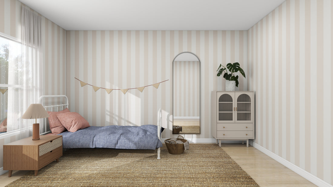 A cozy bedroom with vibrant Circus Stripe Wallpaper Soft Beige by munks and me features a bed adorned with a blue blanket and pink pillows. A wooden nightstand with a lamp sits nearby, while a cabinet holding a plant and mirror is on the opposite wall, accompanied by a floor wicker basket and overhead pennant banner.