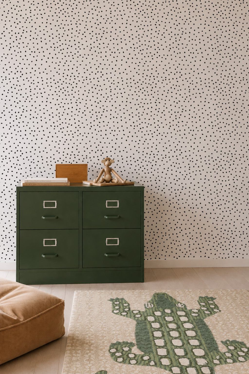 A green four-drawer dresser with a teddy bear and books on top sits against a white wall adorned with the Cora Spotty Wallpaper Sample in Black, adding a playful touch. A beige cushion and a rug featuring a green crocodile design are arranged on the light wooden floor.