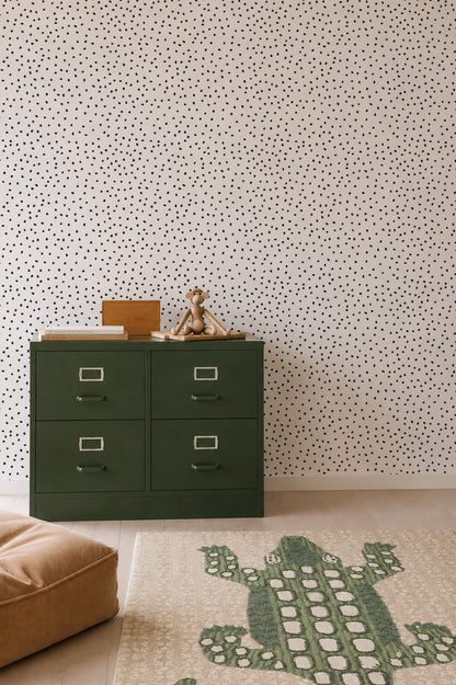 A green four-drawer dresser with a teddy bear and books on top sits against a white wall adorned with the Cora Spotty Wallpaper Sample in Black, adding a playful touch. A beige cushion and a rug featuring a green crocodile design are arranged on the light wooden floor.