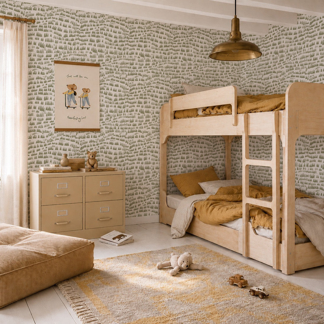 A cozy kids’ bedroom features light wood bunk beds, mustard yellow bedding, a patterned rug, pouf, dresser, and playful Daphne Drops Wallpaper in Olive. Sunlight streams through sheer curtains for a warm, inviting vibe.