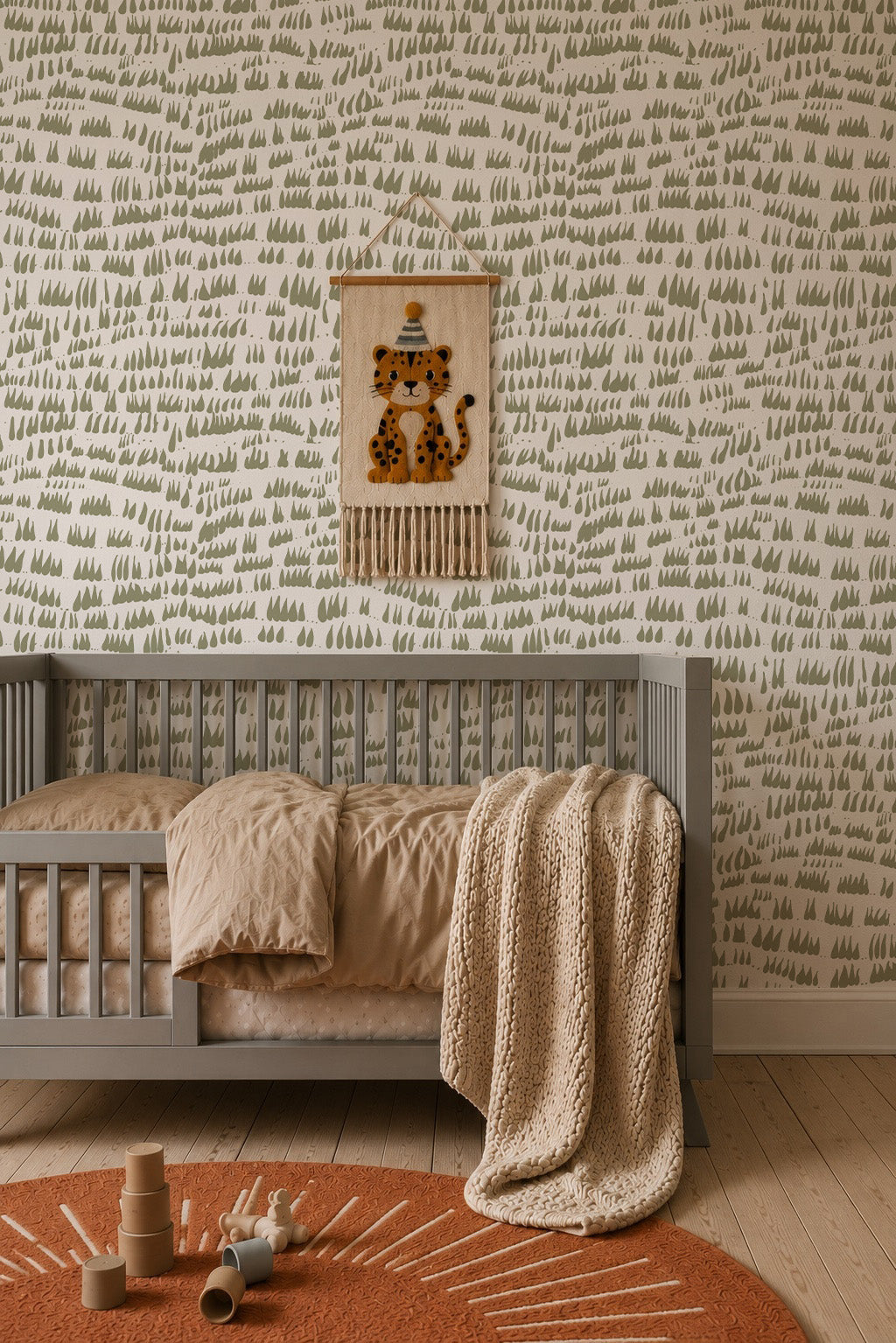 A cozy nursery with a gray crib, tan bedding, chunky knit blanket, and playful animal wall hanging. The walls are lined with Daphne Drops Wallpaper in Olive, while wooden toys rest on a round rust-colored rug.
